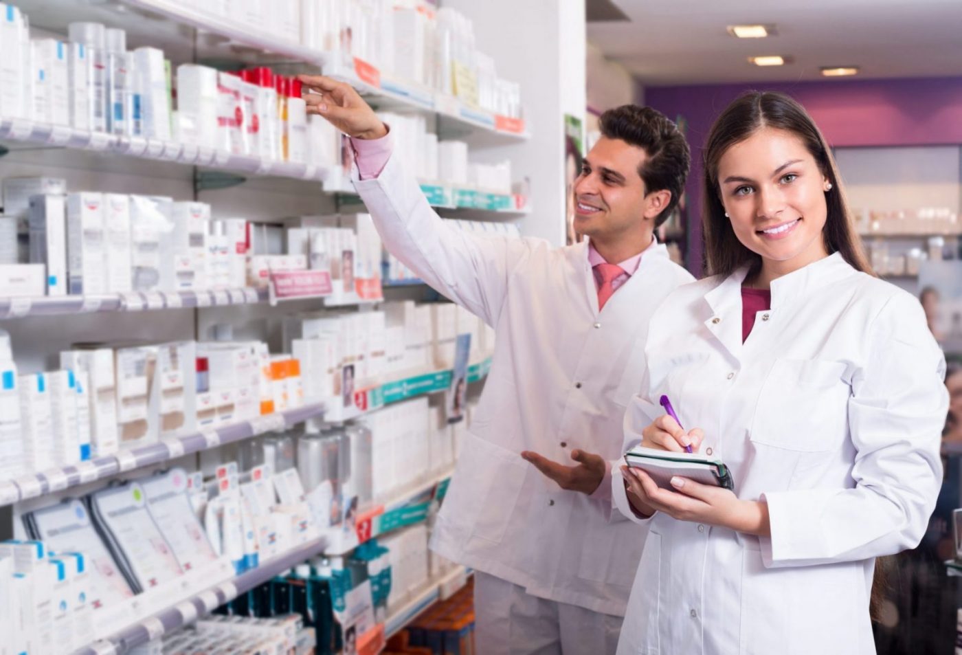 Dos farmacéuticos junto a estanterías de medicamentos, uno toma notas — formación para el empleo