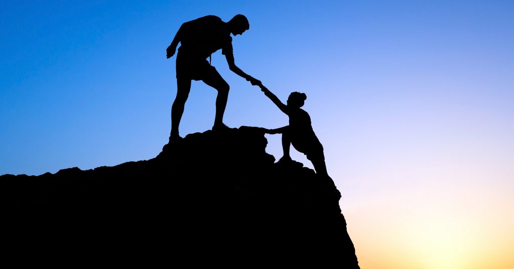 Silueta de dos personas en una roca al atardecer, una ayudando a la otra — formación para el empleo