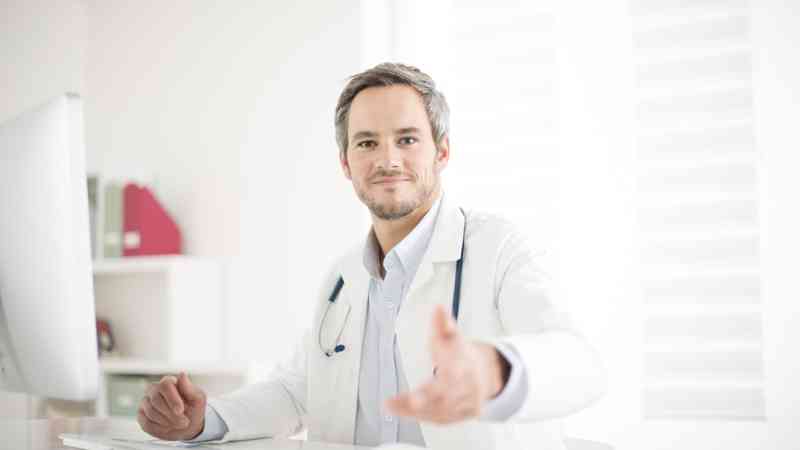 médico con bata y estetoscopio frente al ordenador ofreciendo la mano — cursos online
