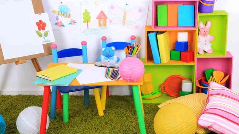 Mesa infantil multicolor con material de manualidades, sillas pequeñas y estantería con juguetes — formación para el empleo