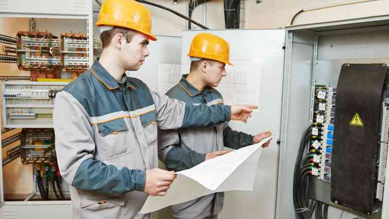 Dos operarios con cascos y uniformes revisan planos junto a un cuadro eléctrico — mantenimiento industrial