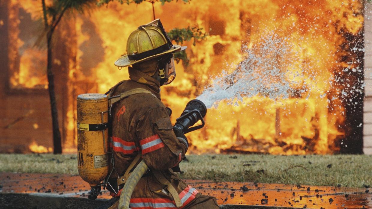 Bombero con equipo y botella de aire rociando agua contra llamas — formación para el empleo