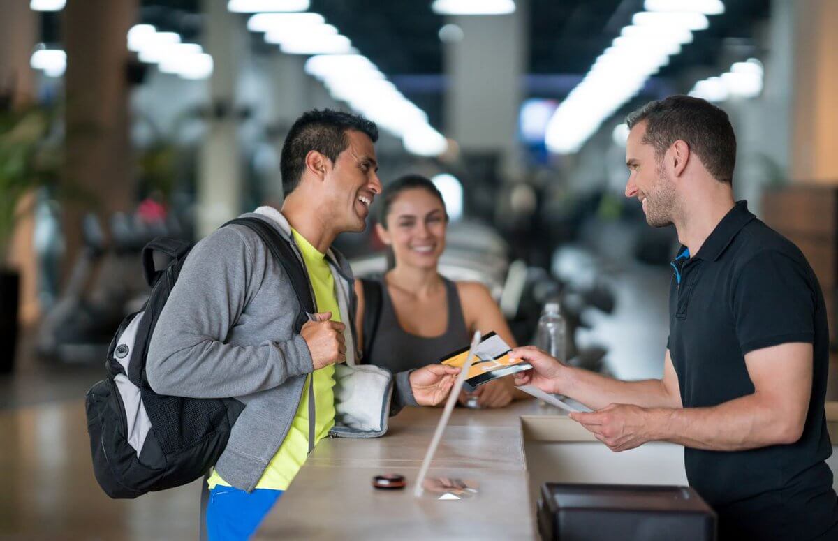 Recepción de gimnasio con empleado entregando tarjeta a dos clientes, formación para el empleo