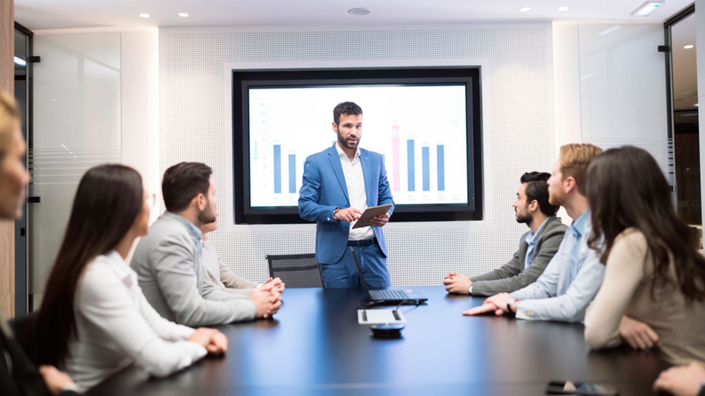 Presentador en traje azul frente a pantalla con gráficos en reunión — formación para el empleo