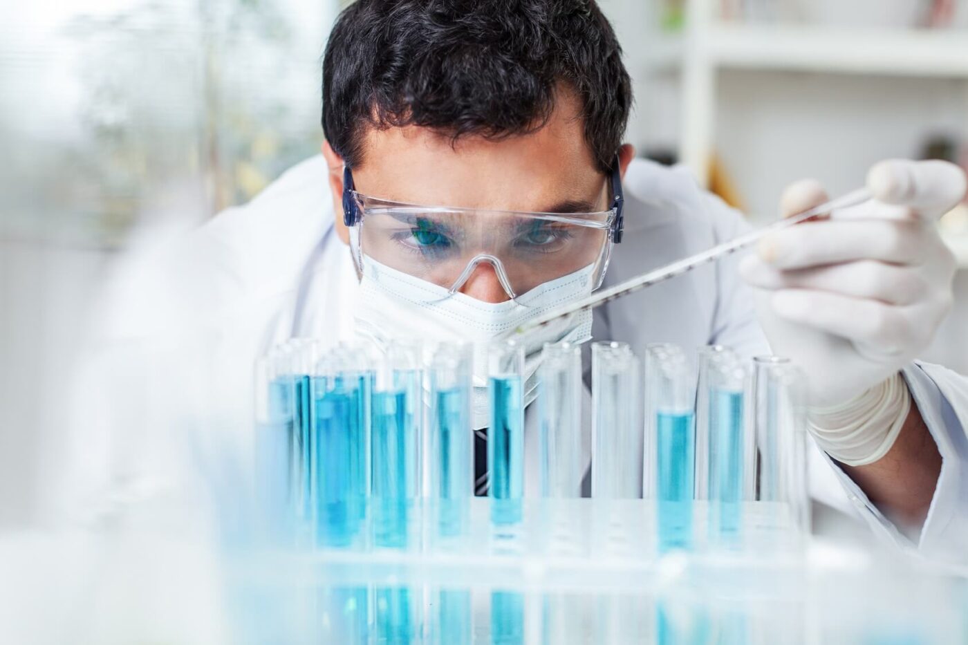 Técnico de laboratorio con gafas, mascarilla y guantes usando una pipeta sobre tubos de ensayo con líquido azul — formación para el empleo