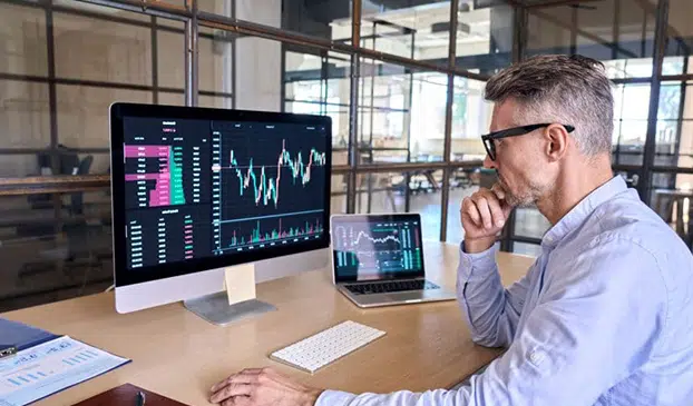 Hombre con gafas mirando gráficos financieros en monitor y portátil — cursos online