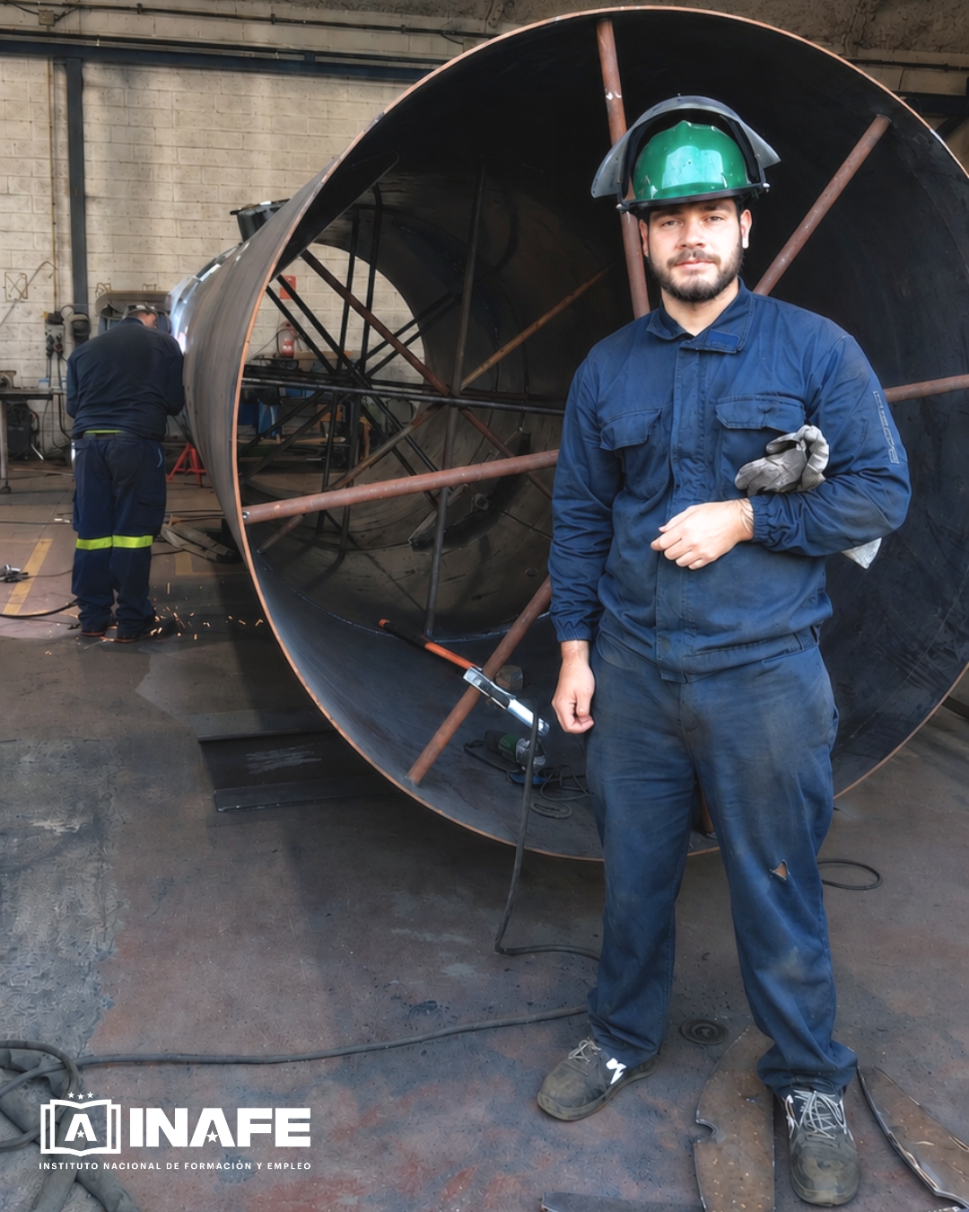 Operario con casco verde junto a una pieza cilíndrica metálica en taller, soldadura