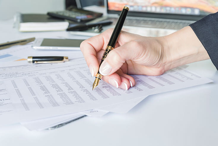 Mano sosteniendo un bolígrafo sobre hojas con tablas numéricas junto a un portátil, cursos online