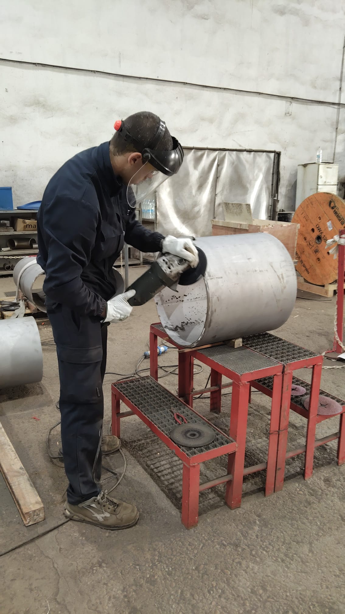 Operario con careta y guantes desbastando un cilindro metálico con amoladora, calderería industrial
