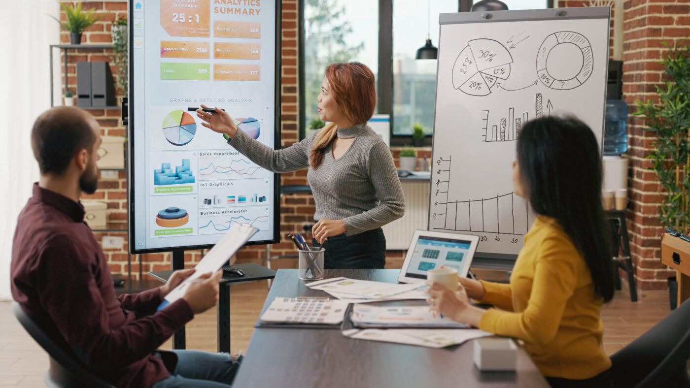 Presentadora señalando gráficos en pantalla durante formación para el empleo, con dos colegas observando.