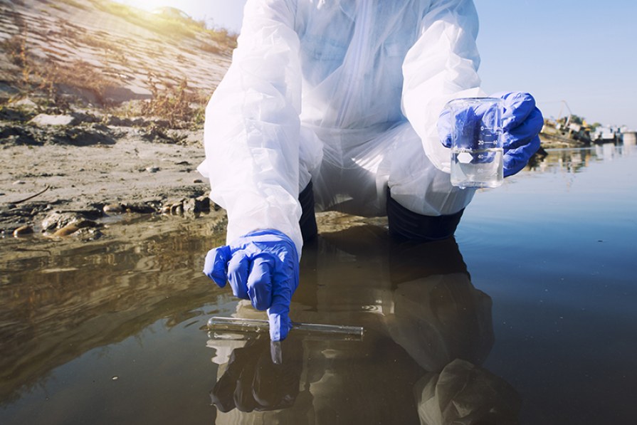 Toma de muestra de agua en la orilla por técnico con traje protector y guantes azules, cursos sector industrial