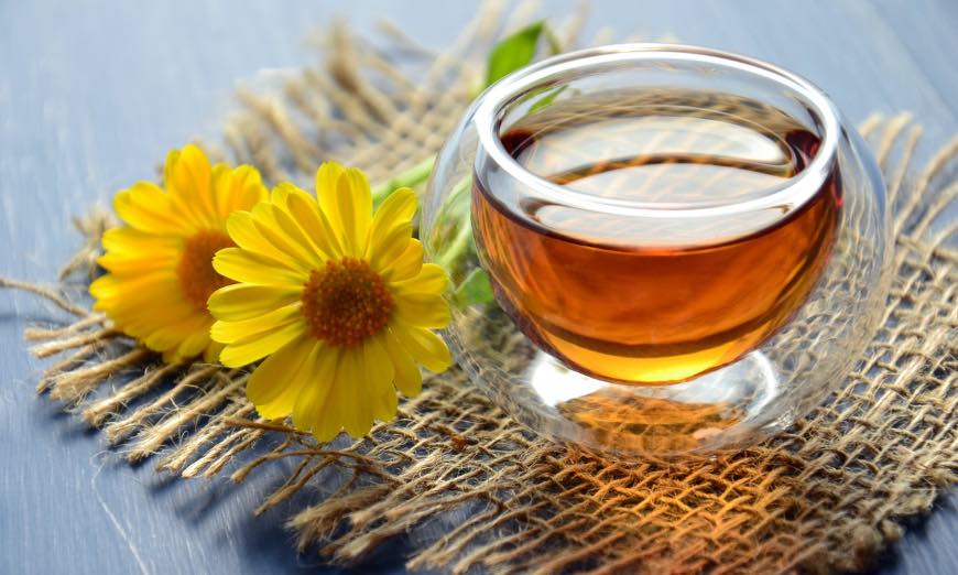 Taza de infusión de manzanilla en cristal con flores amarillas sobre arpillera, cursos online