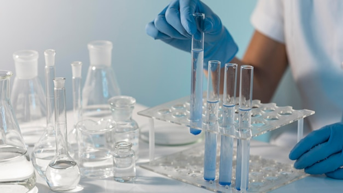 Manos con guantes azules sosteniendo tubos de ensayo con líquido azul en una gradilla, formación para el empleo