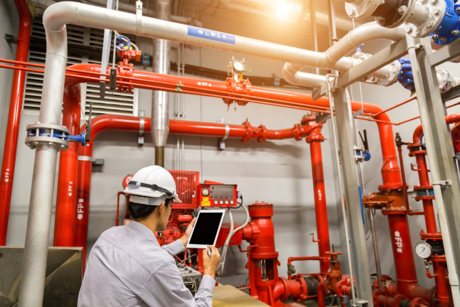 Técnico con casco y tablet inspeccionando bombas y tuberías rojas de un sistema contra incendios, mantenimiento industrial