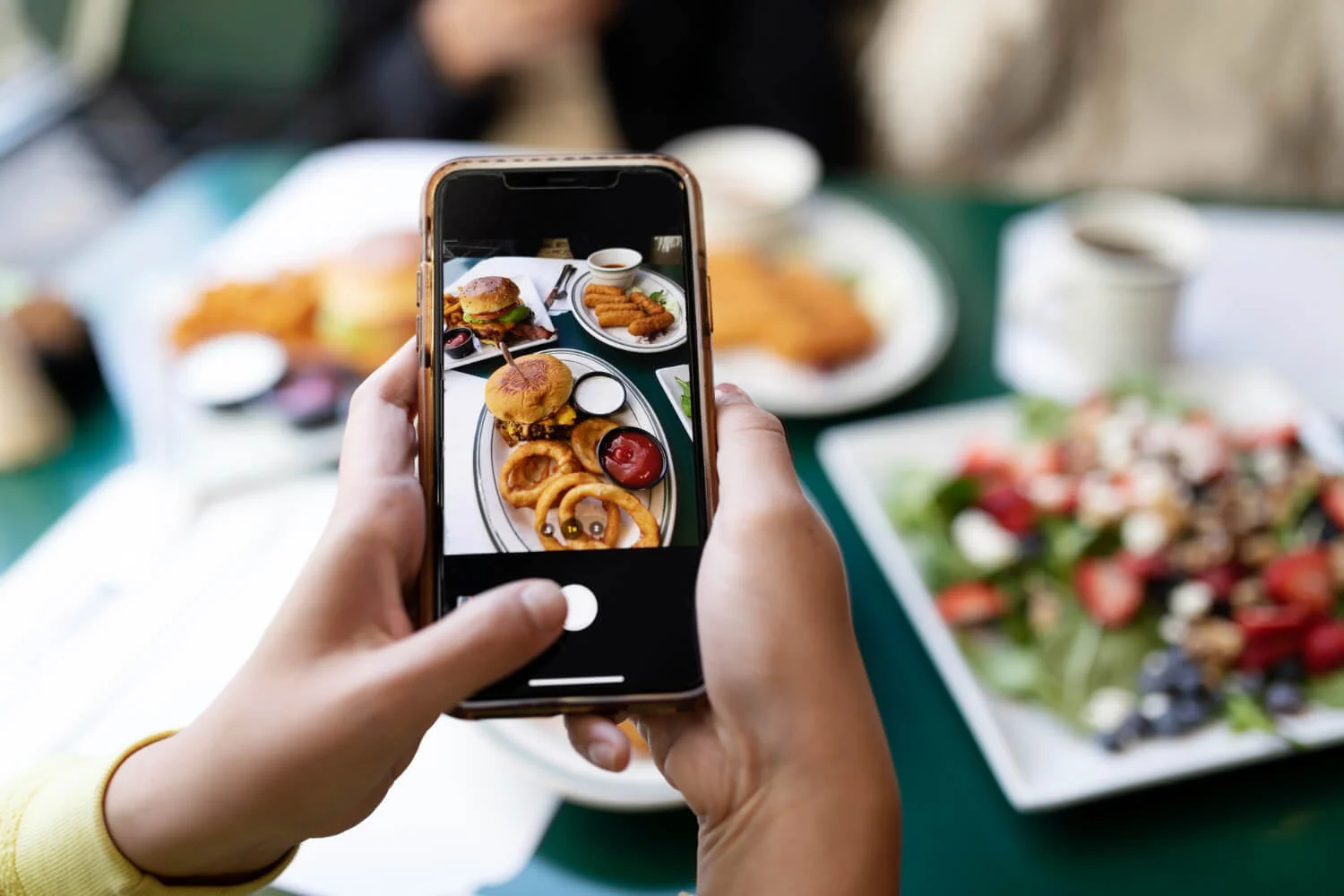 Manos sosteniendo un móvil fotografiando una hamburguesa con aros de cebolla (cursos online)