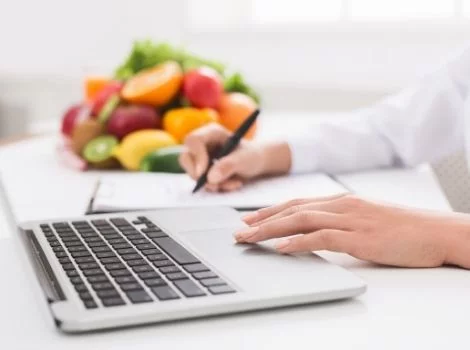 Manos en portátil y escribiendo en papel con cuenco de frutas al fondo (cursos online)