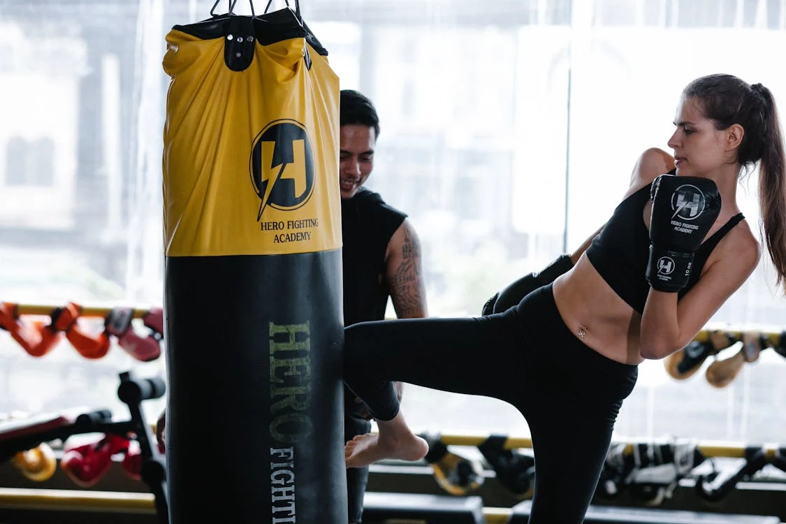 Mujer pateando saco de boxeo amarillo y negro con guantes mientras un entrenador observa — formación a medida