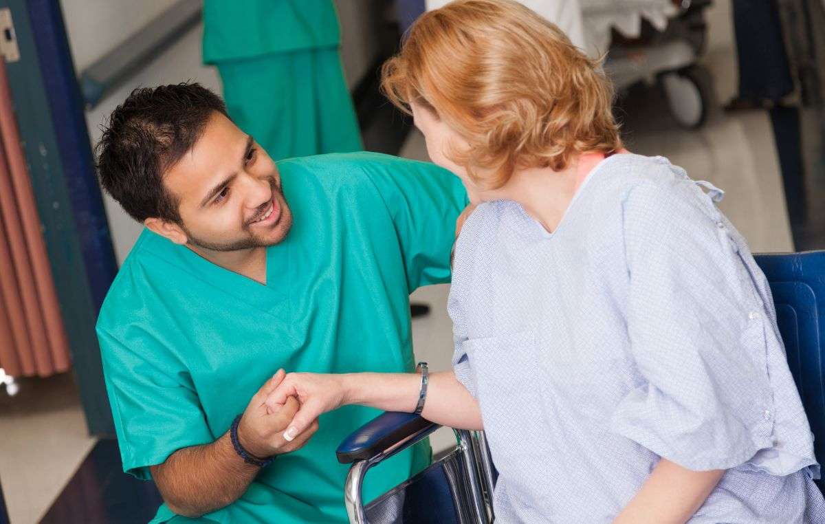 Auxiliar sanitario sonriente sujetando la mano de una paciente en silla de ruedas, certificado de profesionalidad
