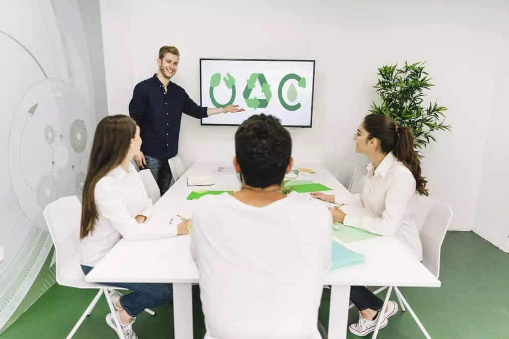 Presentador y grupo en sala con pantalla que muestra símbolos de reciclaje — formación para el empleo