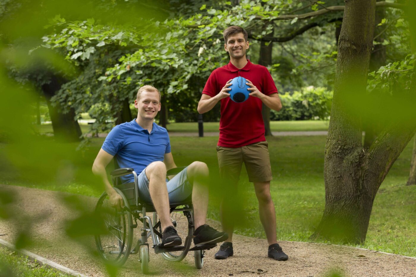 Hombre en silla de ruedas sonriendo junto a un amigo de pie con un balón azul en un parque, formación para el empleo