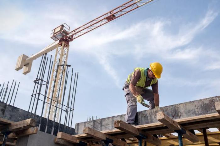 Obrero de construcción con casco y chaleco trabajando sobre un encofrado de madera, grúa de torre al fondo, cursos sector industrial