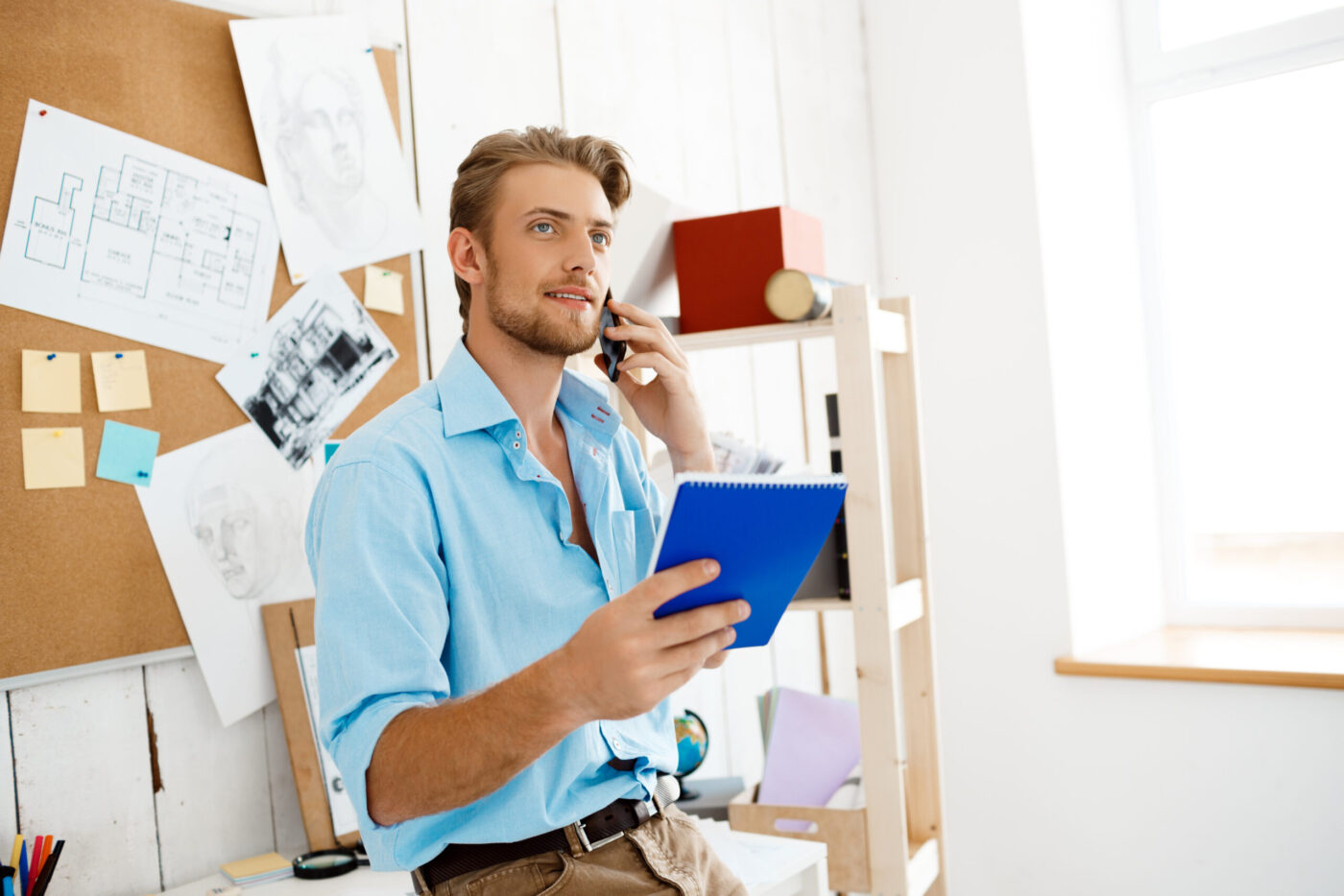 Hombre joven con camisa azul hablando por teléfono y sosteniendo un cuaderno azul, cursos online
